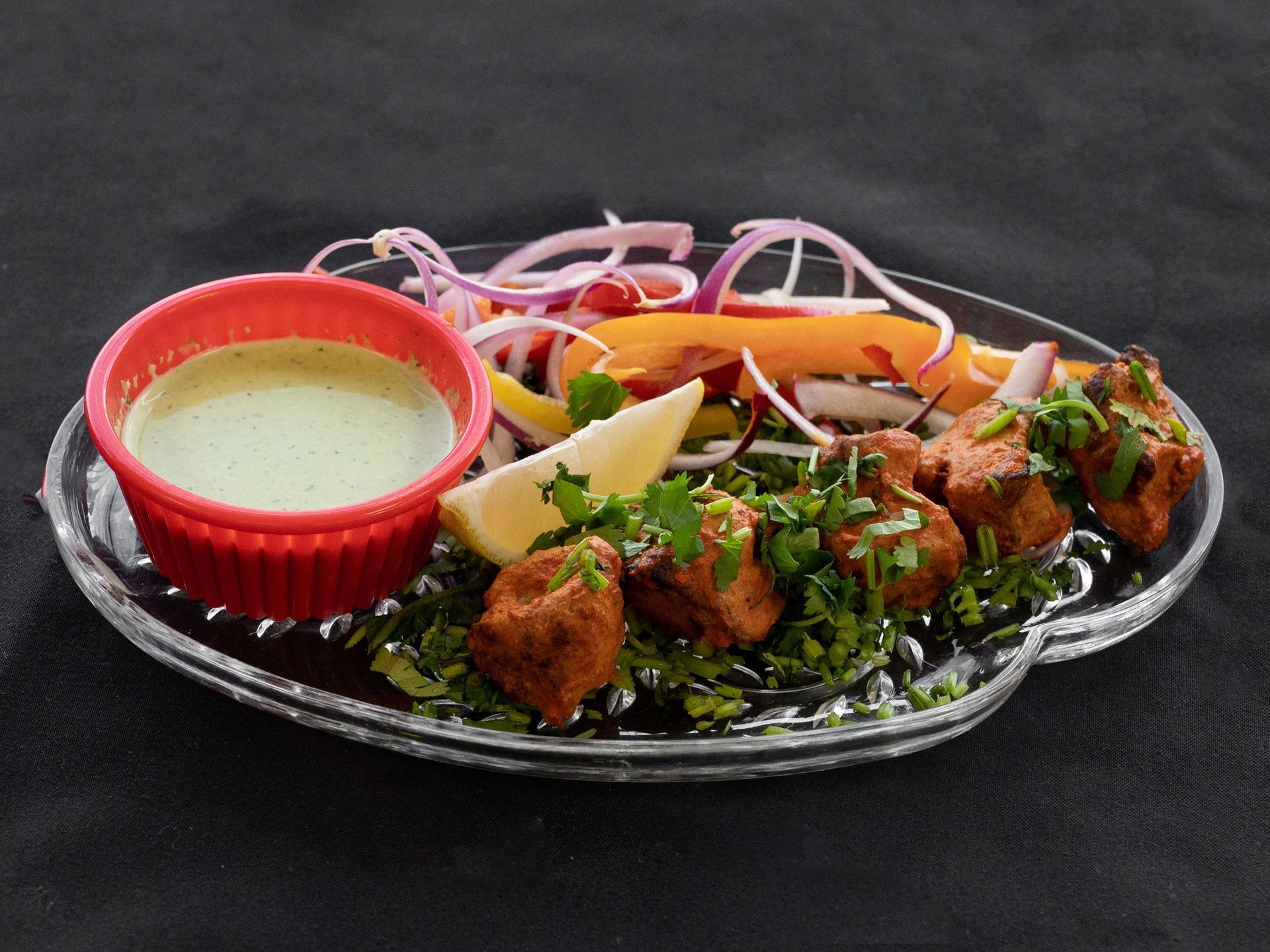Plate of Indian food: Tandoori chicken, mint sauce, salad, lemon, on black surface.