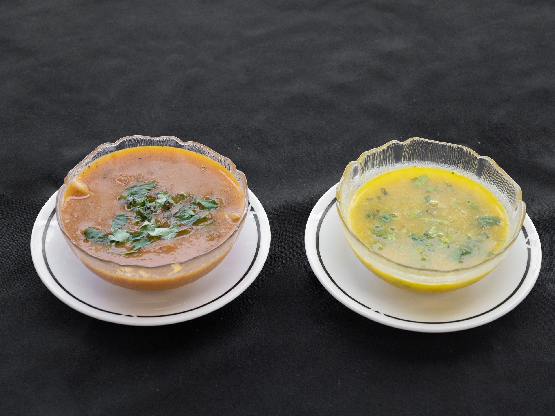 Two bowls of soup on white plates, one brown with green garnish, one yellow with green herbs.