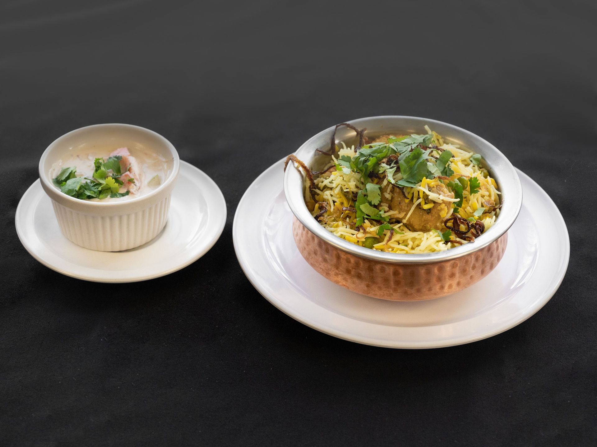 Biryani in copper bowl with raita on a white plate, dark background.