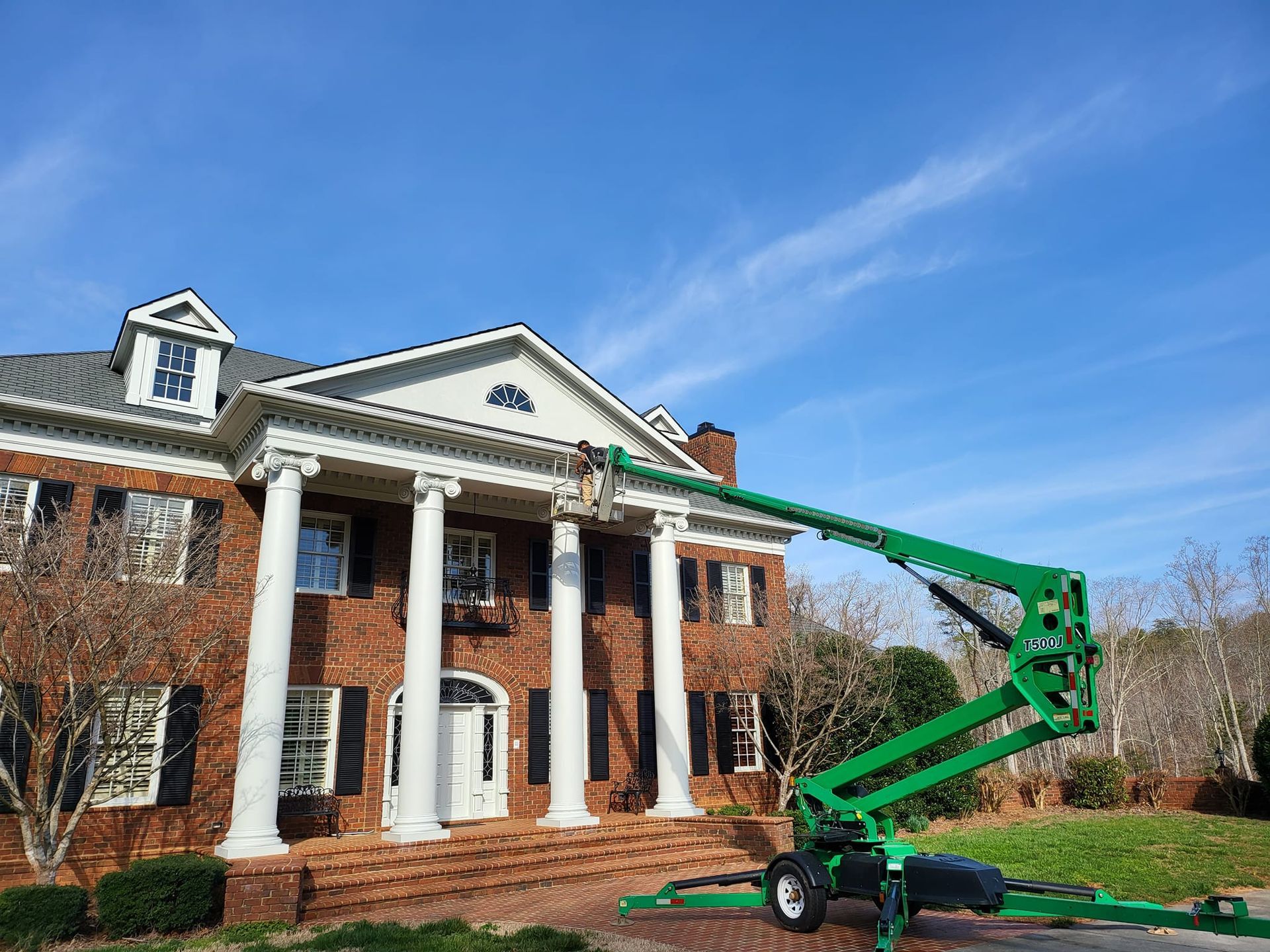 Green lift near a large brick house with white columns, a person working at the roof under a blue sky.
