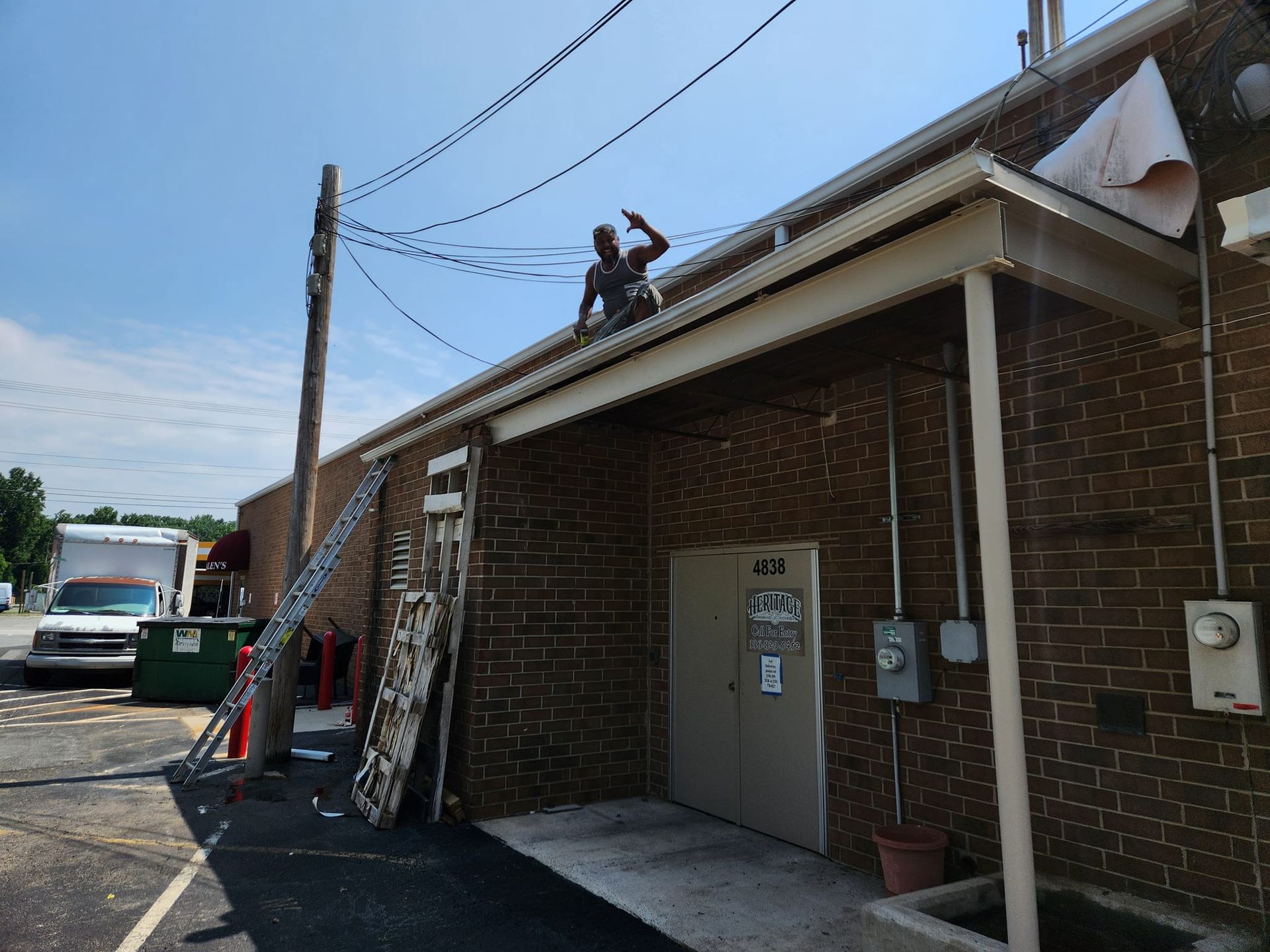 Person on roof near power lines, working on building's gutters. Ladder leaned against brick wall. Sunny day.