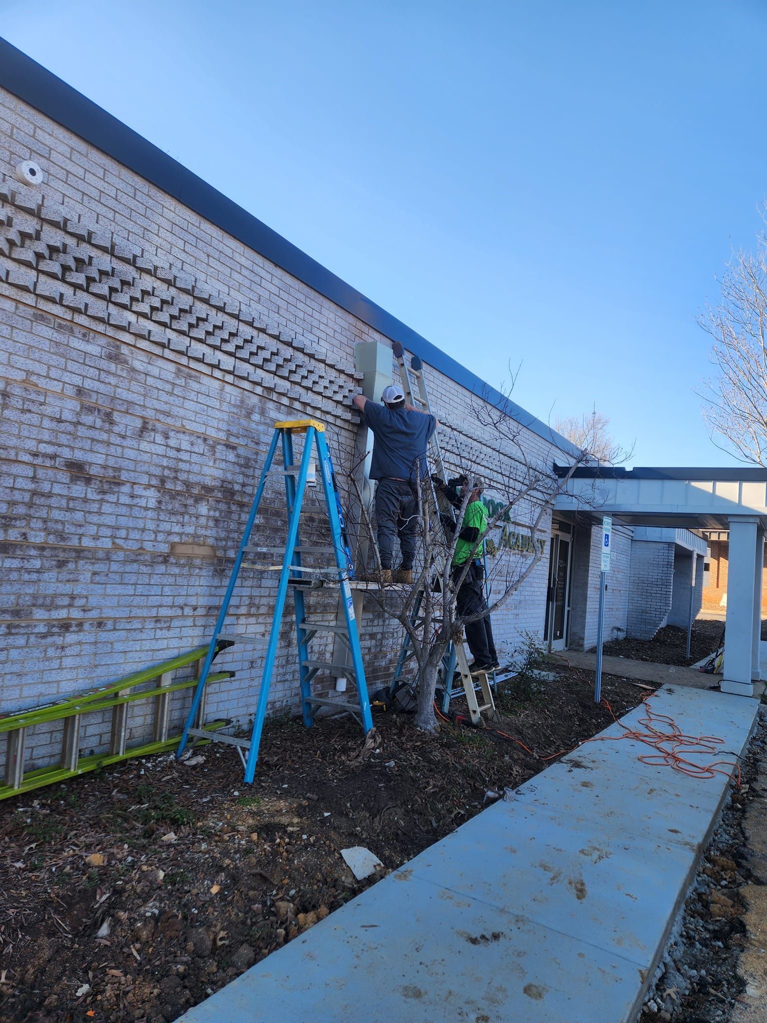 Two workers on ladders installing equipment on a brick building's exterior.