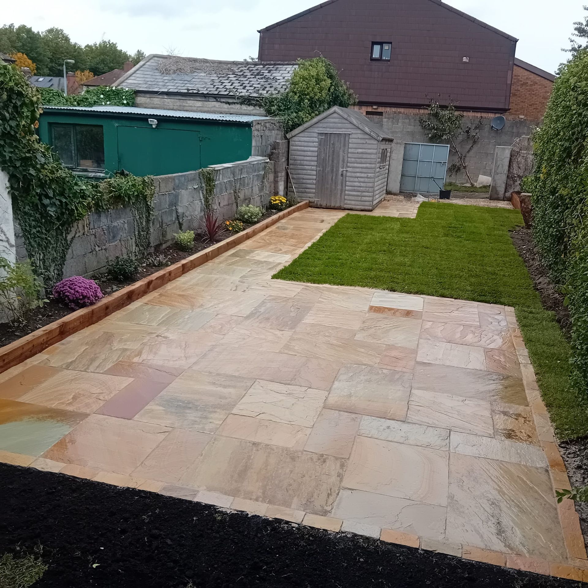 Backyard patio with sandstone pavers, small lawn, and shed. Stone walls and a hedge line the sides.