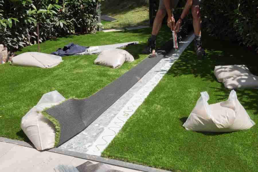 Person installing artificial turf on a patio, with bags of sand and rolls of turf nearby.