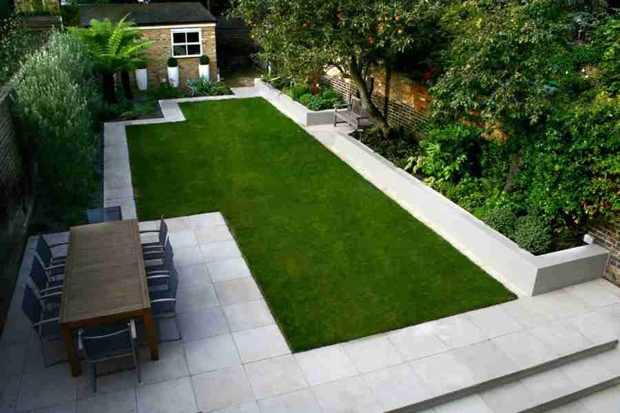Modern garden with a green lawn, stone patio, and dining table, surrounded by trees and raised beds.