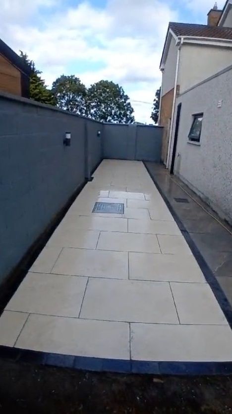A paved outdoor walkway with cream-colored tiles, framed by a dark border and gray walls.