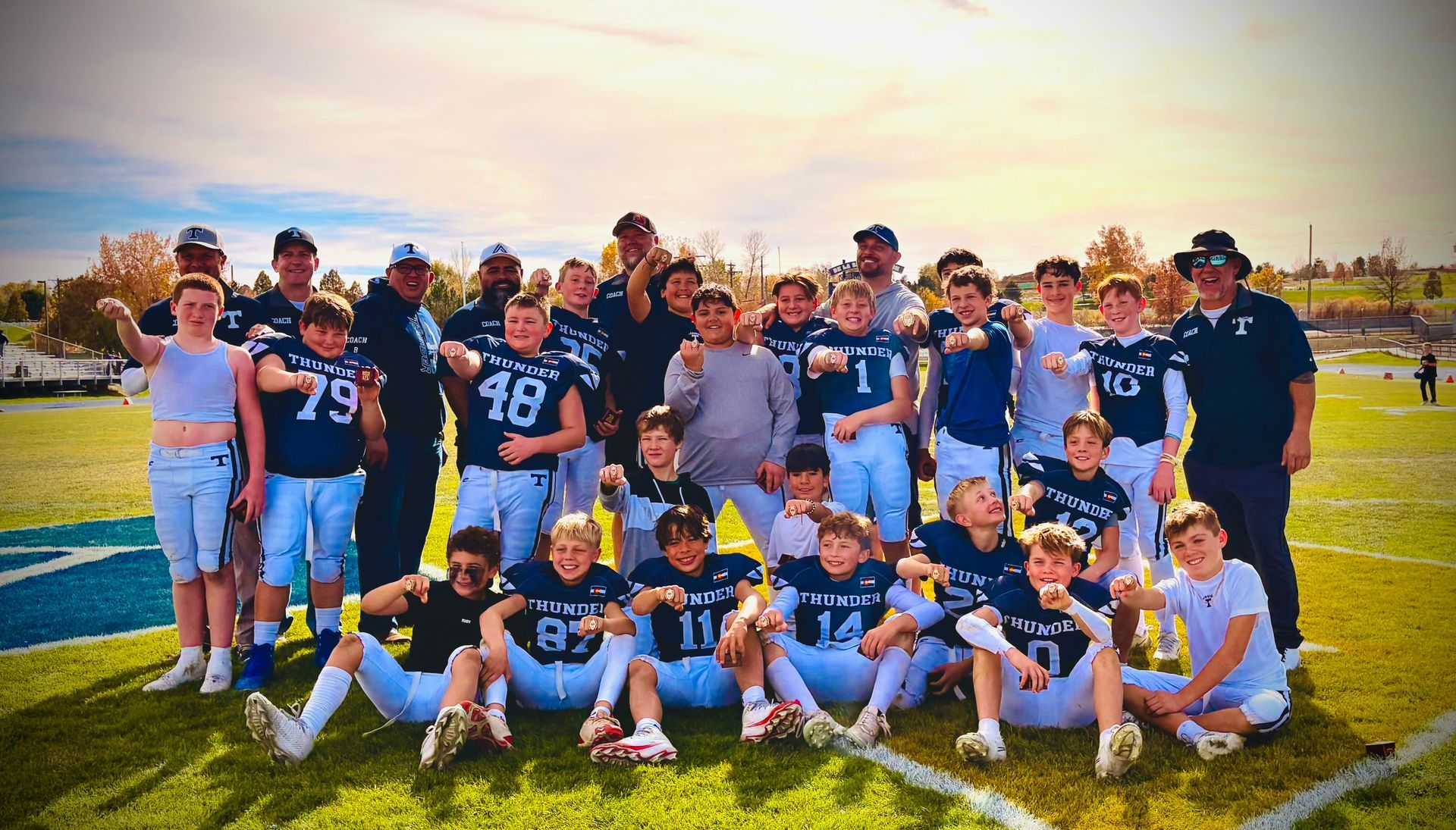 Youth football team posing on a field at sunset, wearing navy and white uniforms with trophies in front.
