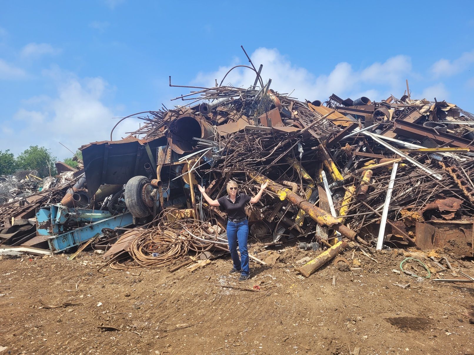 Woman stands before a large pile of scrap metal under a blue sky, arms raised.