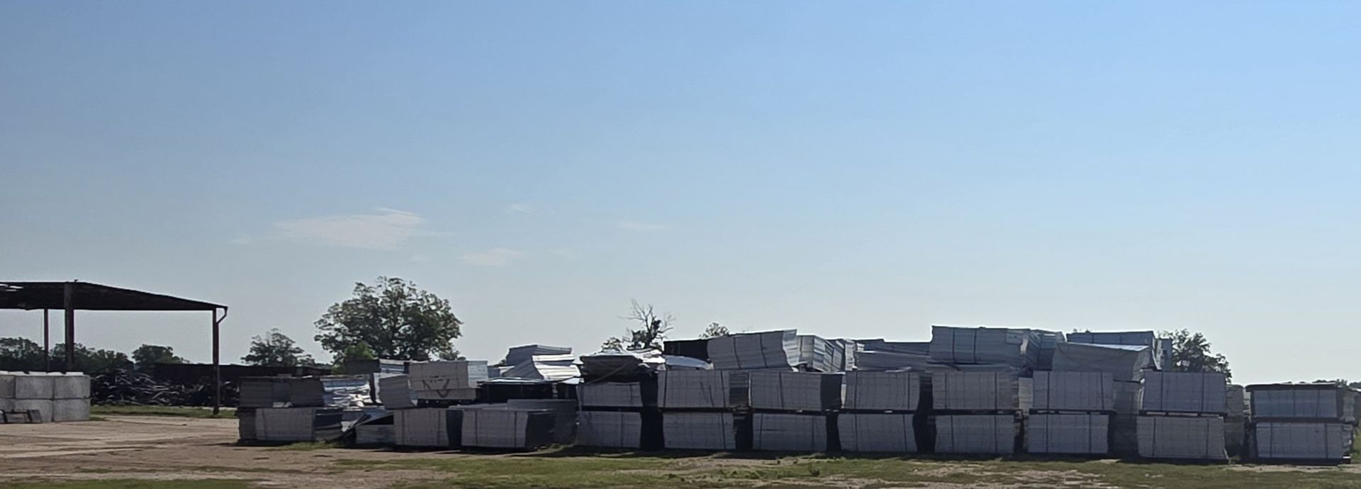 Piles of construction materials are stacked outside on a grassy area, under a blue sky.