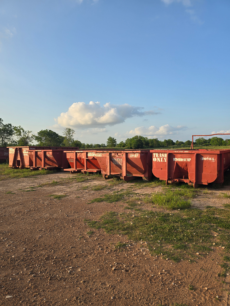Red metal bins lined up outdoors under a blue sky, next to trees and grass.