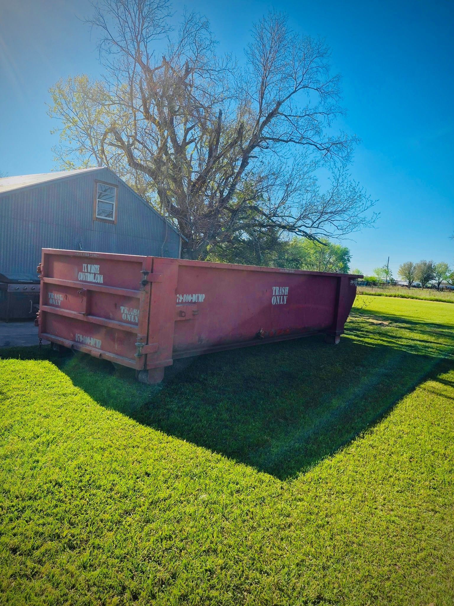 Red dumpster on green grass, next to a barn, under a blue sky.