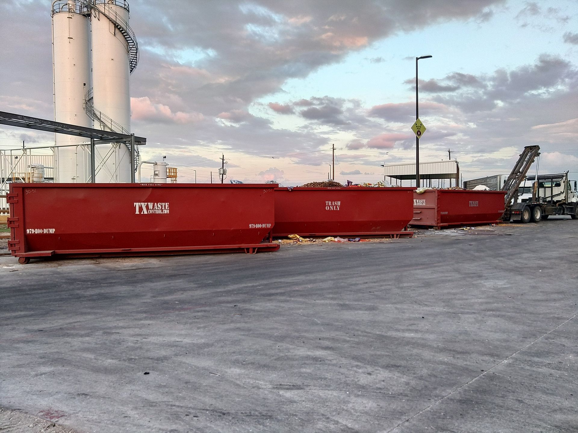 Red industrial dumpsters lined up outdoors with a truck, silos, and a cloudy sky.