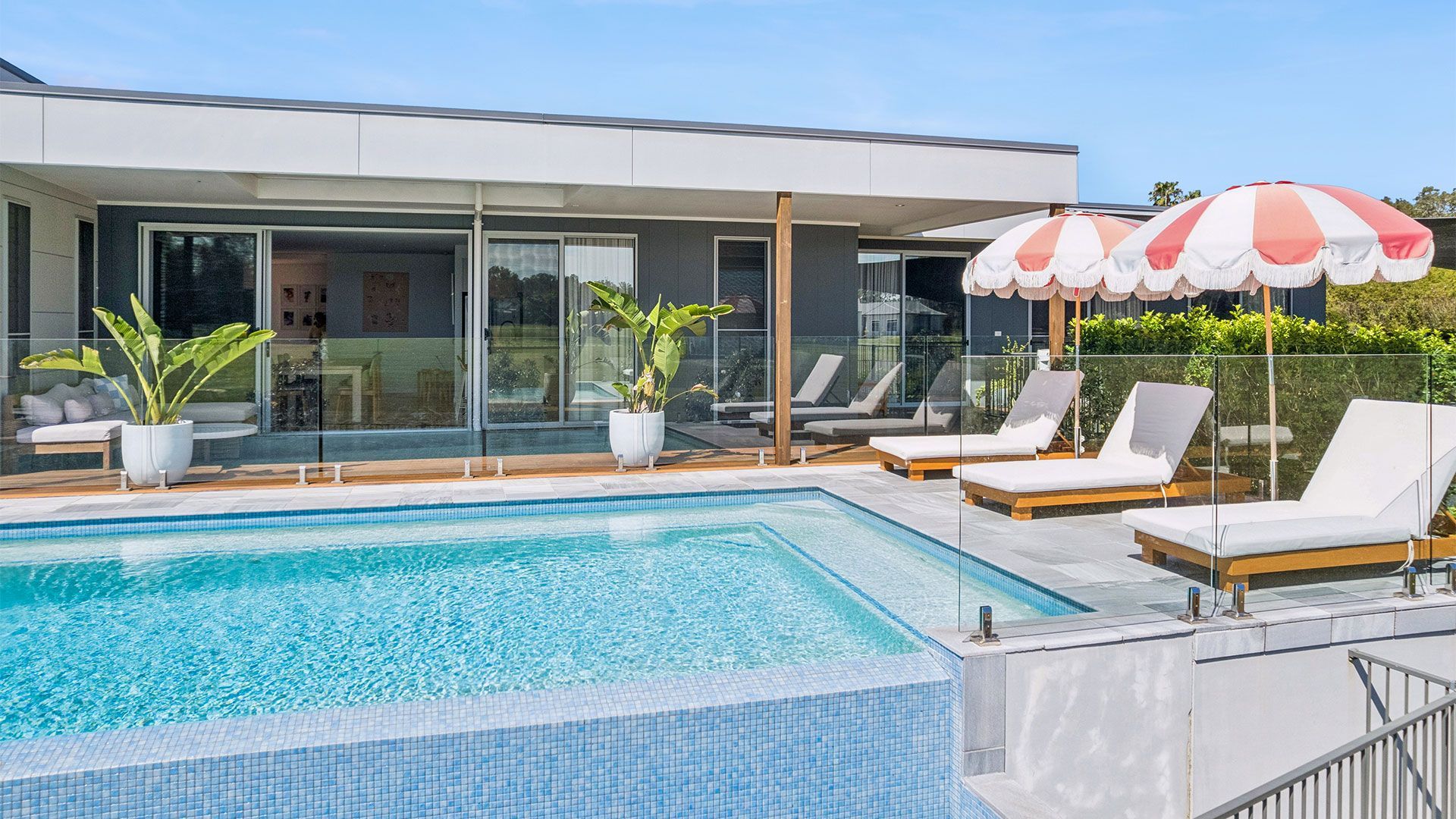 Modern backyard pool with lounge chairs, striped umbrella, and a glass-walled house in the background