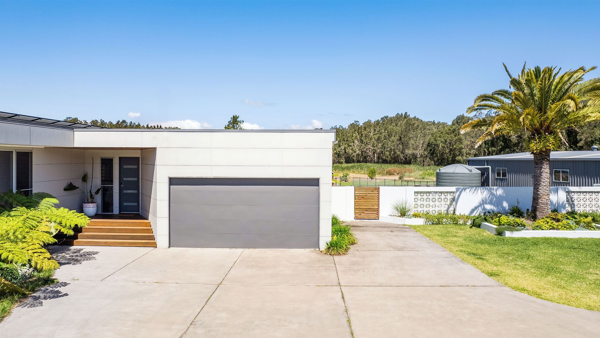 Modern white house with a gray garage door, driveway, lawn, and palm tree under a blue sky