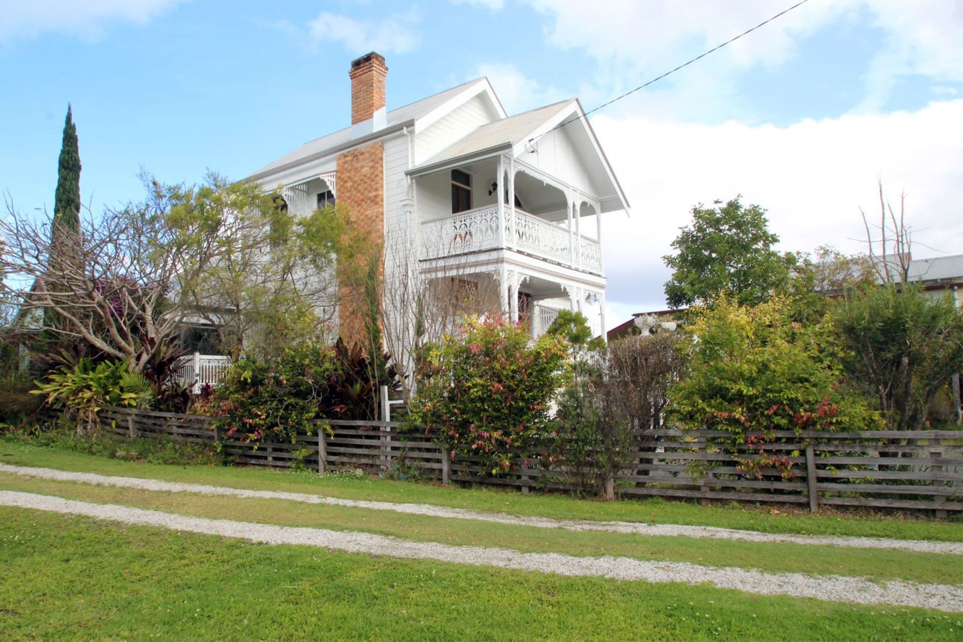 White two-story house with a porch, surrounded by greenery and a wooden fence beside a grassy road.