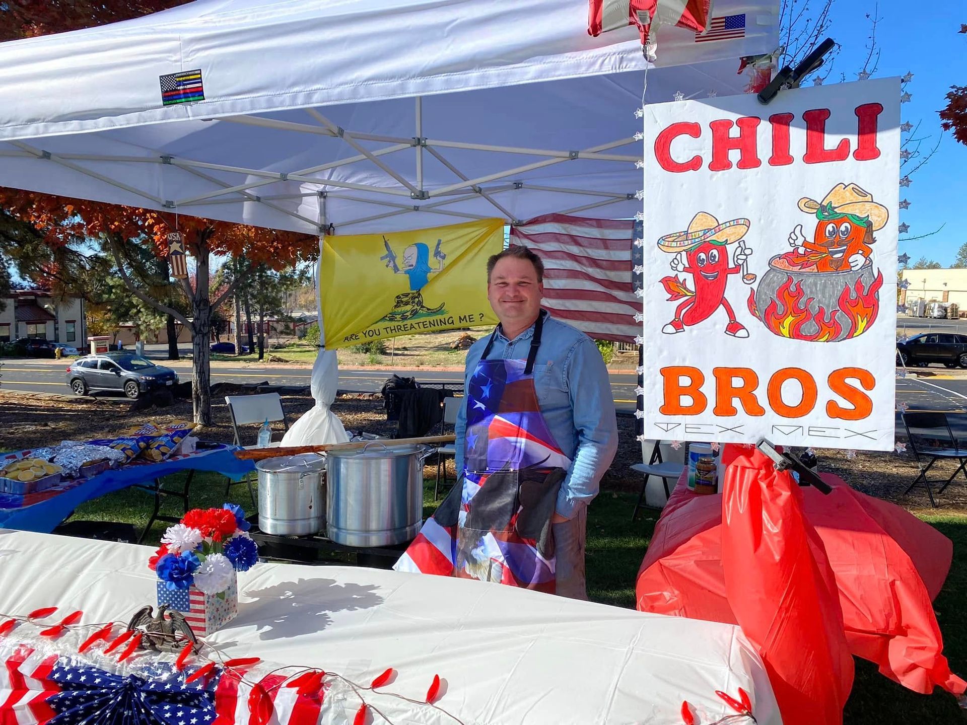 Man behind chili stand sign reading “CHILI BROS” at an outdoor market, with red-white-blue table display