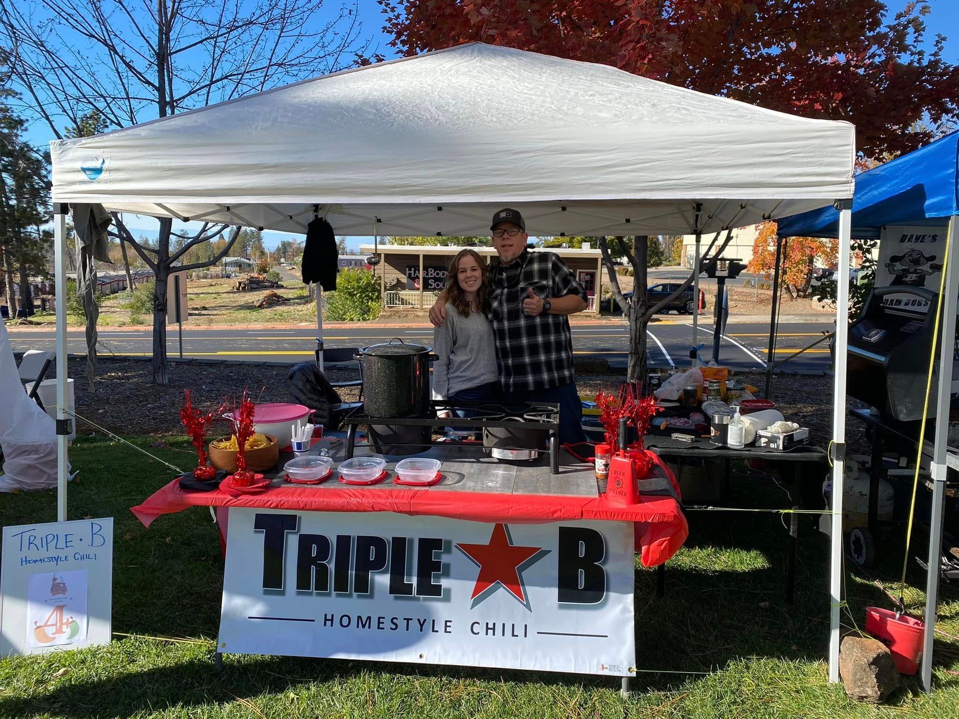 Outdoor vendor booth under a white canopy with a TRIPLE B banner, two people standing behind the table.
