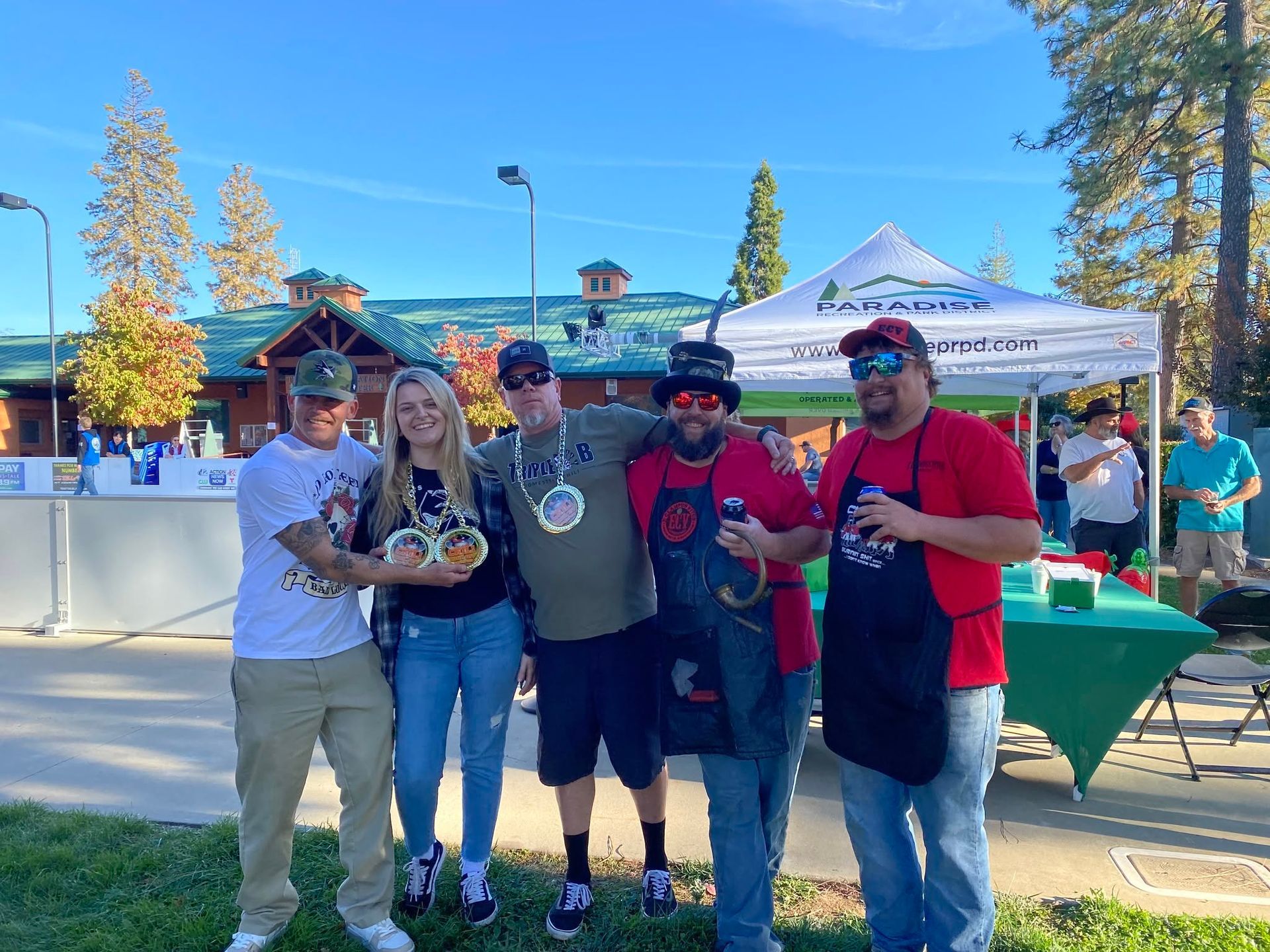 Five people posing with drinks at an outdoor event under a canopy on a sunny day