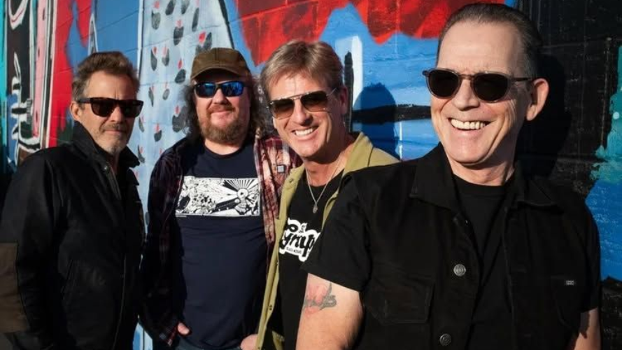Four smiling people posing in sunglasses before a colorful mural wall