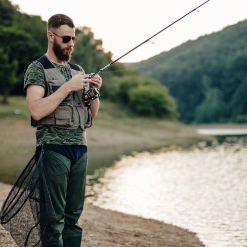 Man fishing on a lake shore, holding a fishing rod with a net at his side.