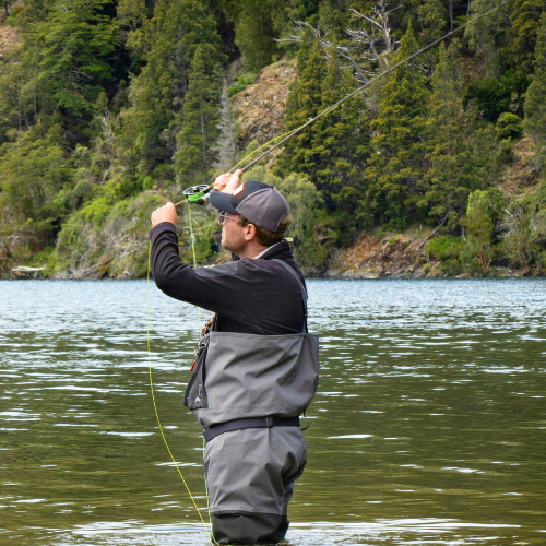 Person fly fishing in a river beside a forested rocky shoreline.