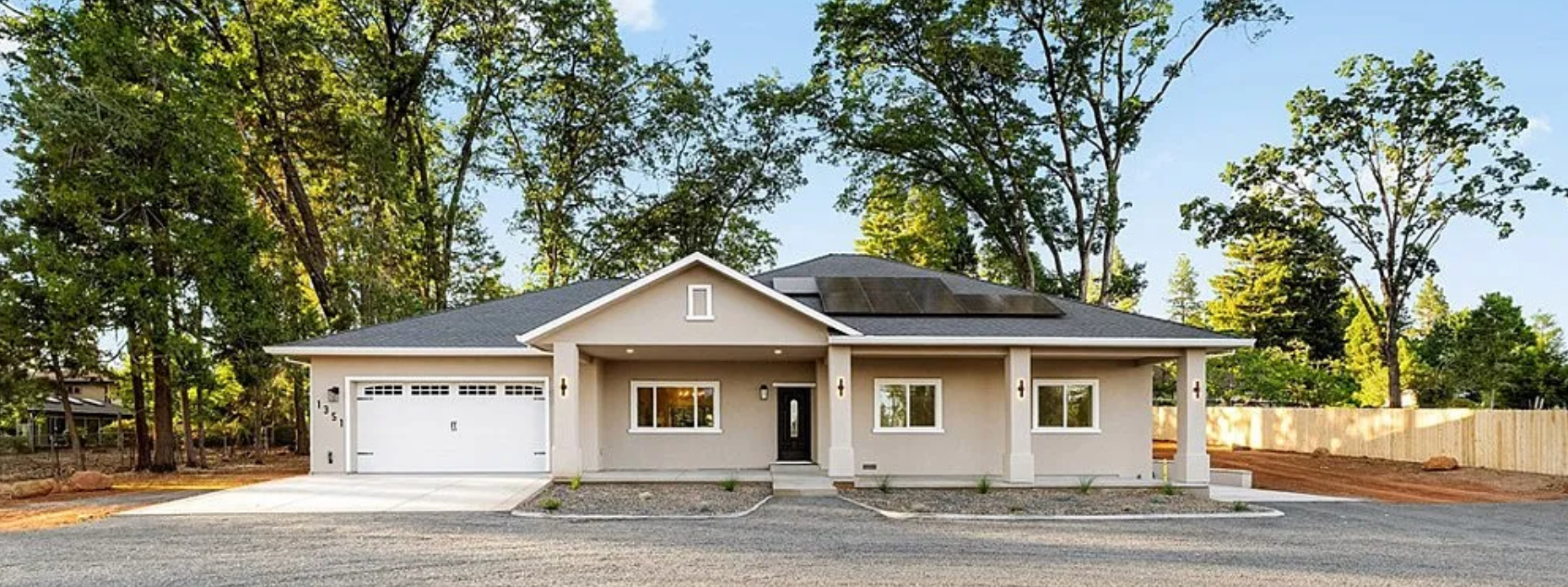 Single-story beige house with white trim, front porch, and attached garage on a wooded lot.