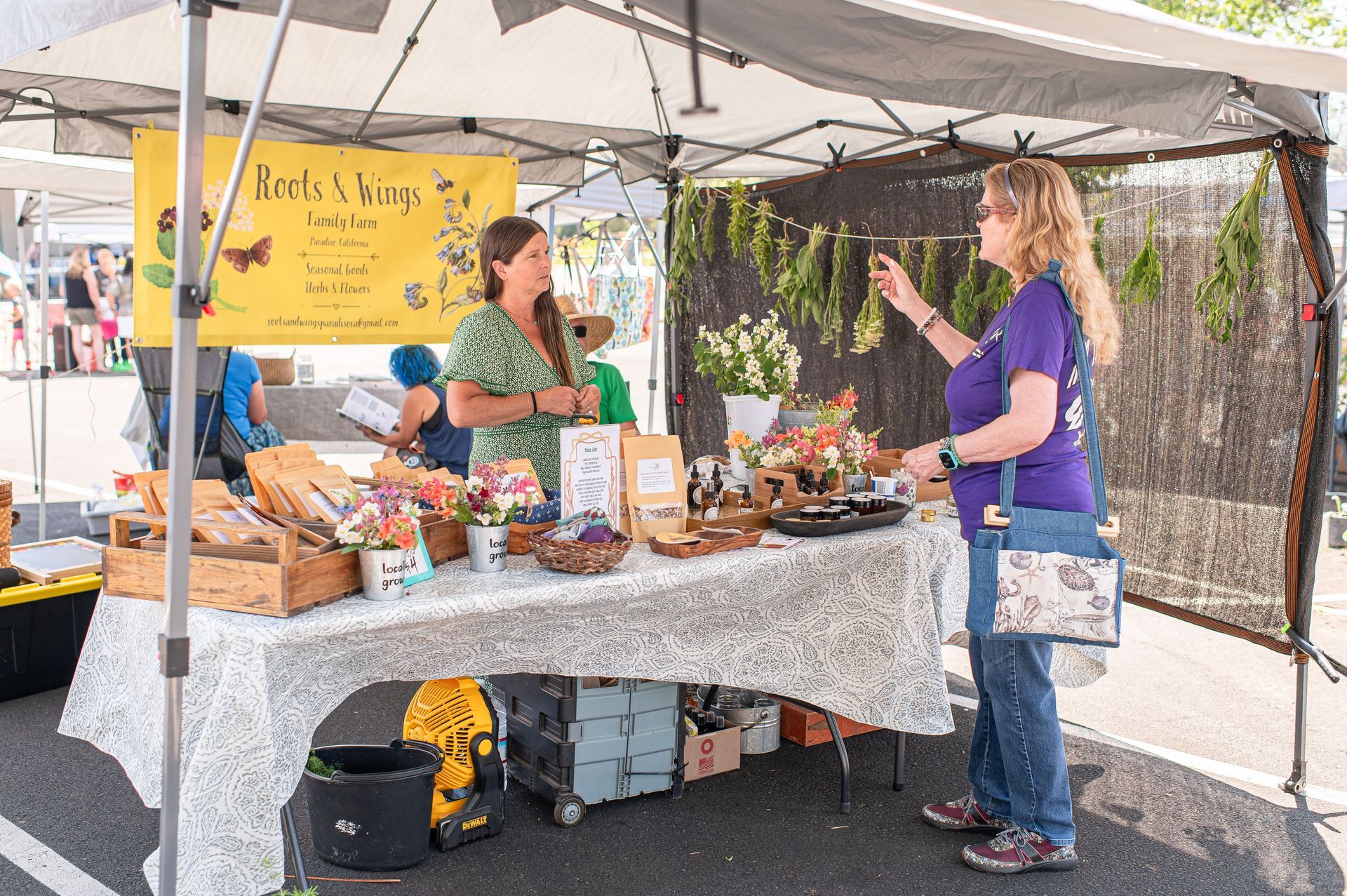 Two women talking at an outdoor market stall under a canopy, with baked goods and jars on display.