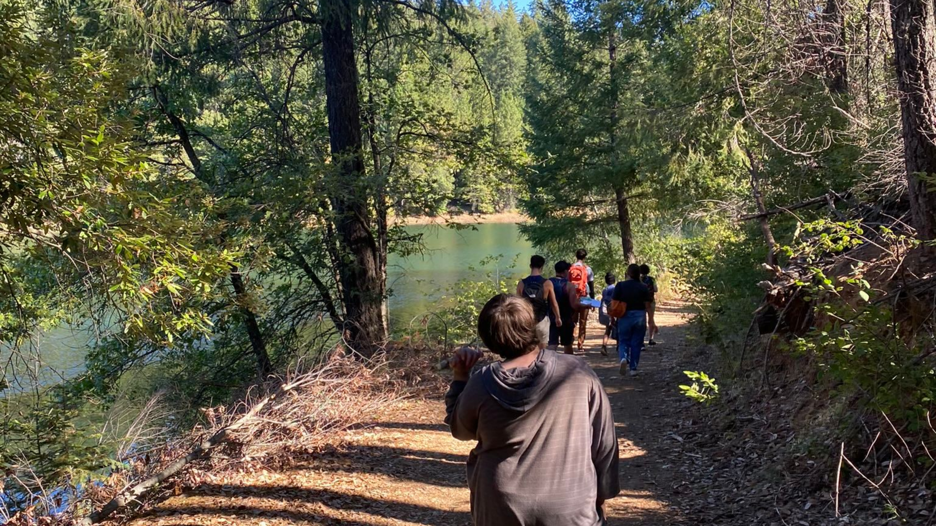 People walking along a wooded riverside trail in sunlight, with trees and water nearby.