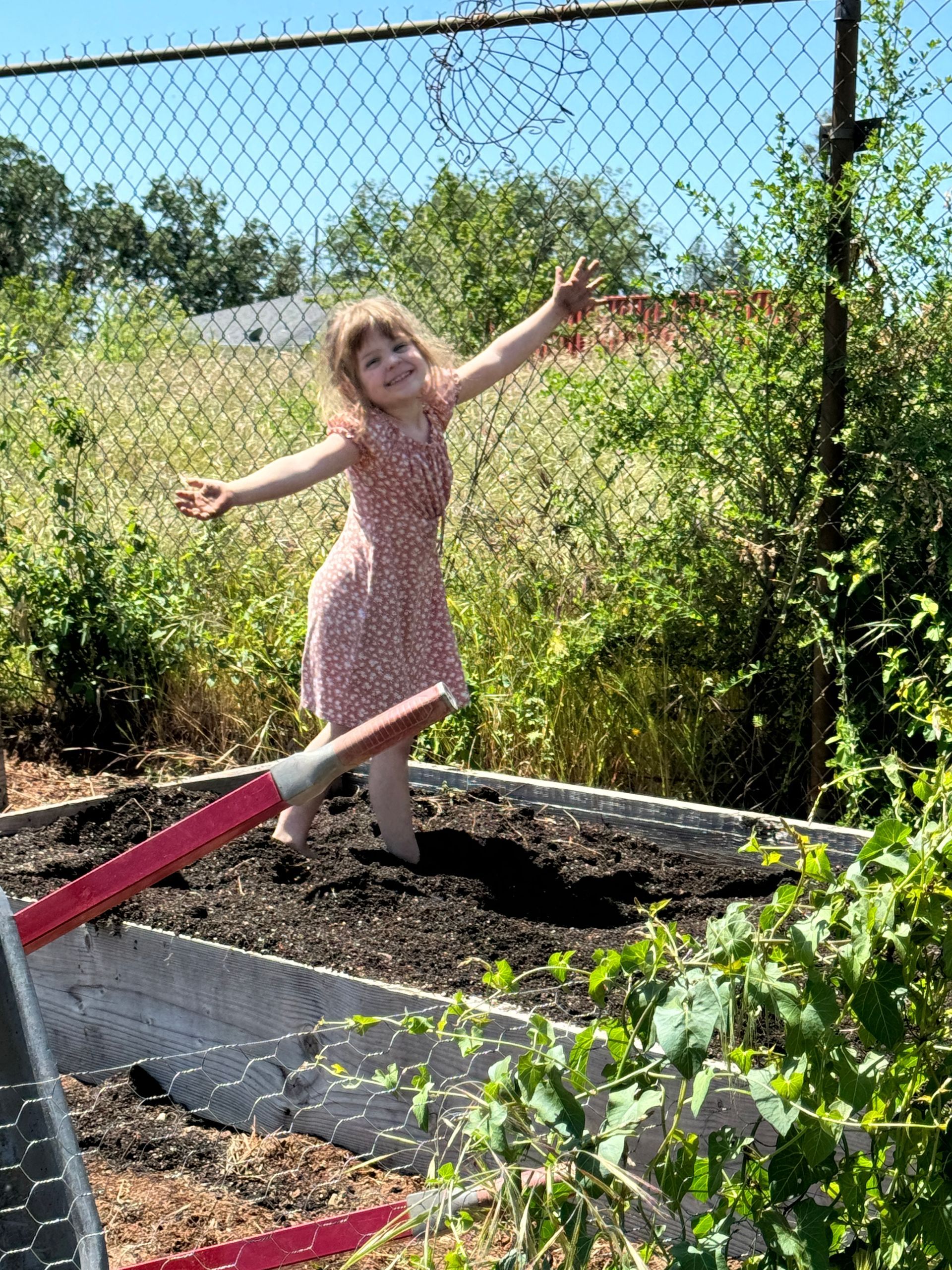 Child in a pink dress balancing on a red beam in a sunny garden beside a chain-link fence.