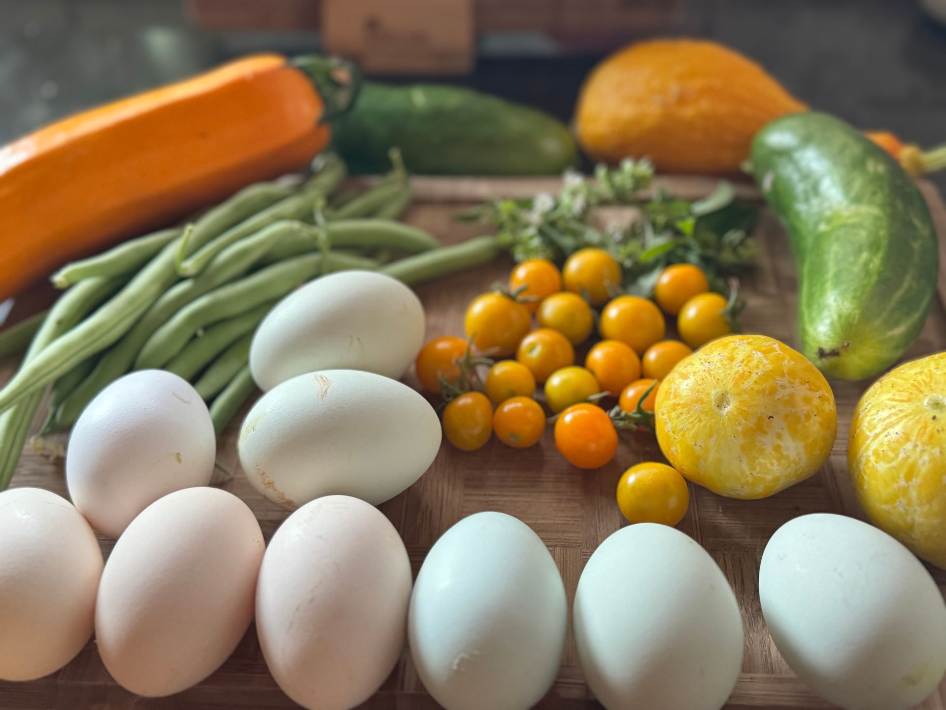 Assorted fresh vegetables and eggs arranged on a table, including green beans, cucumbers, squash, tomatoes, and peppers