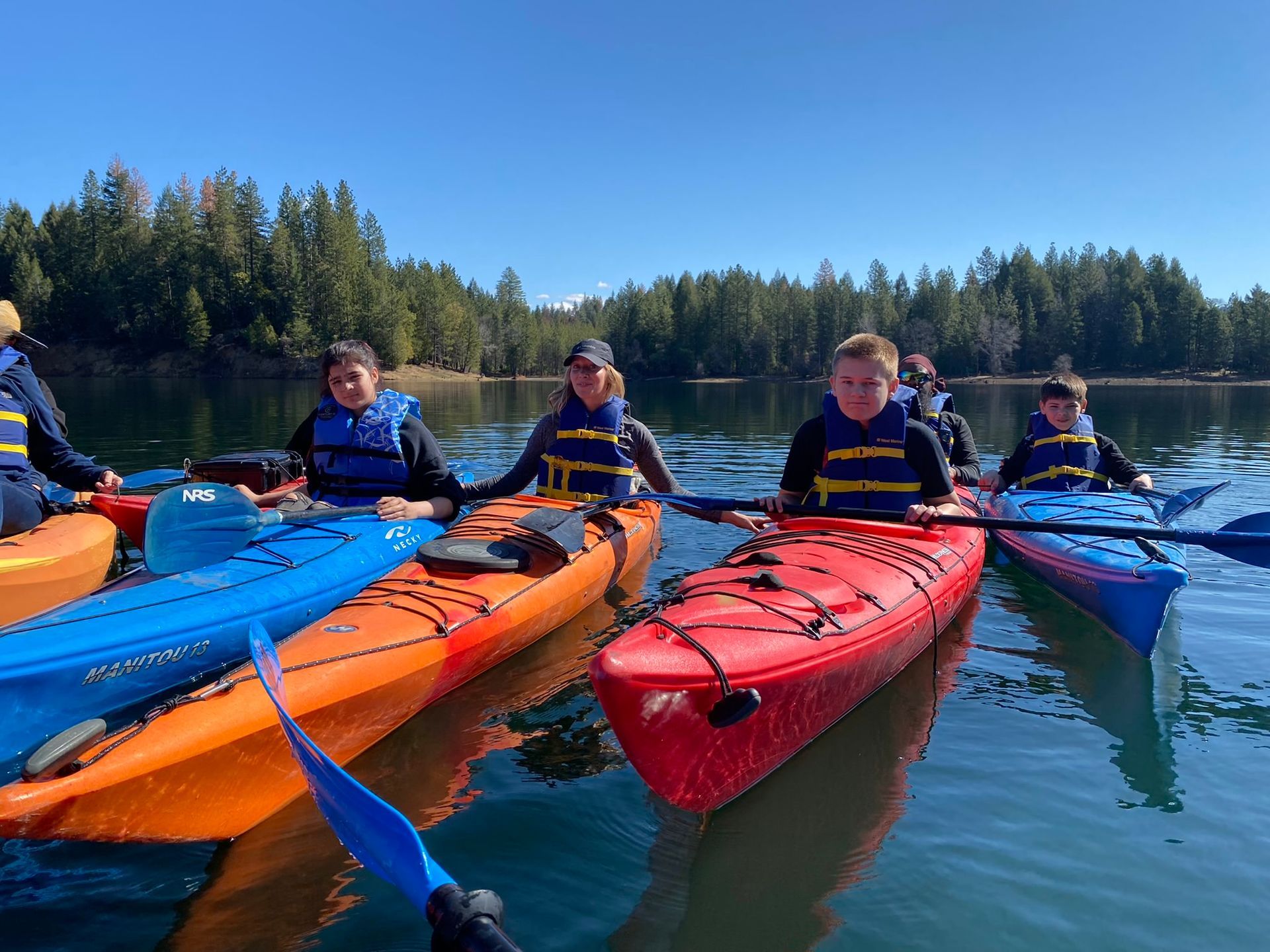 Kayakers on calm water under a blue sky, wearing life vests. Trees line the distant shore.