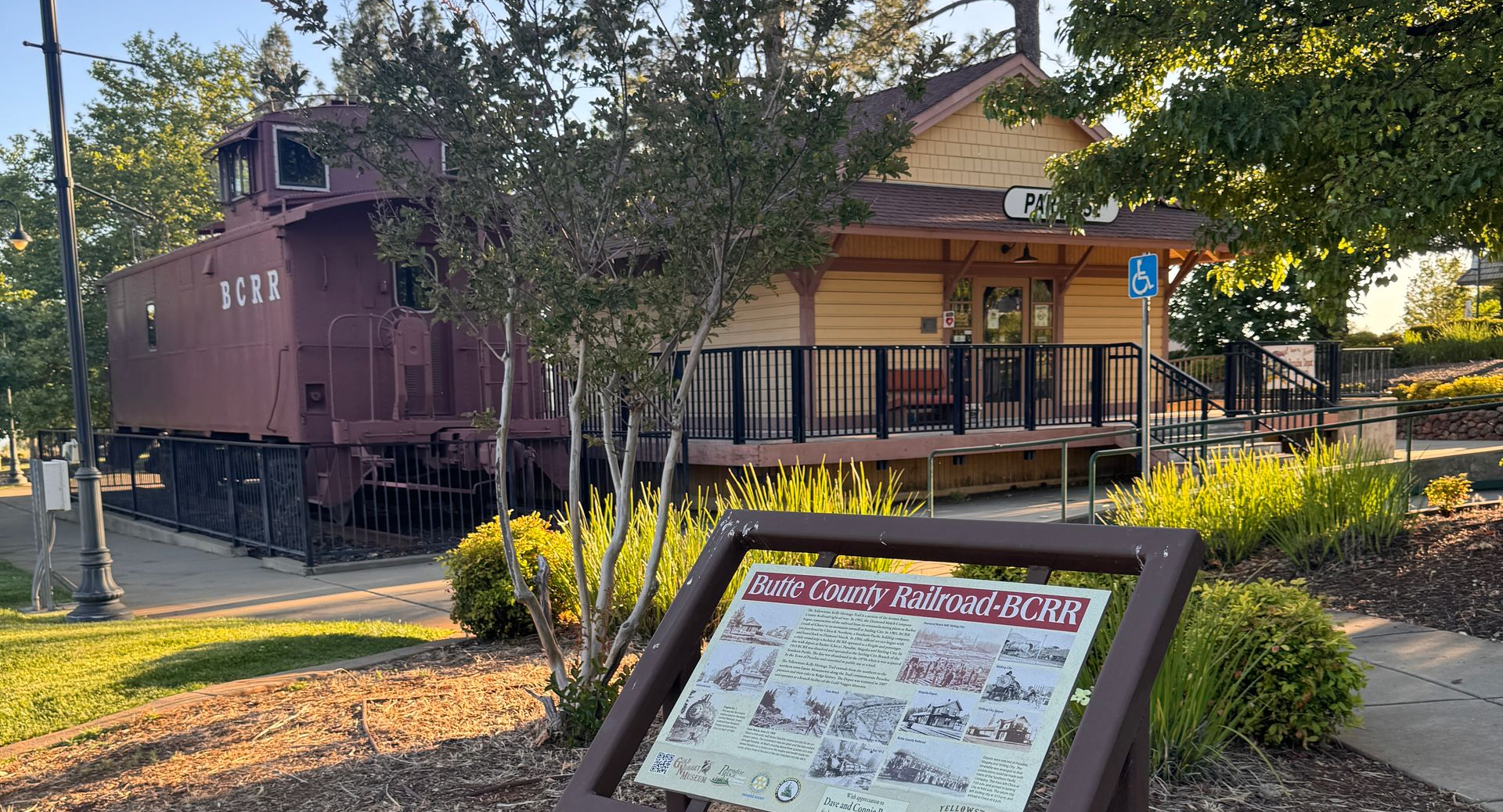 Historic train car and station building in a sunny park, with a sign in the foreground.