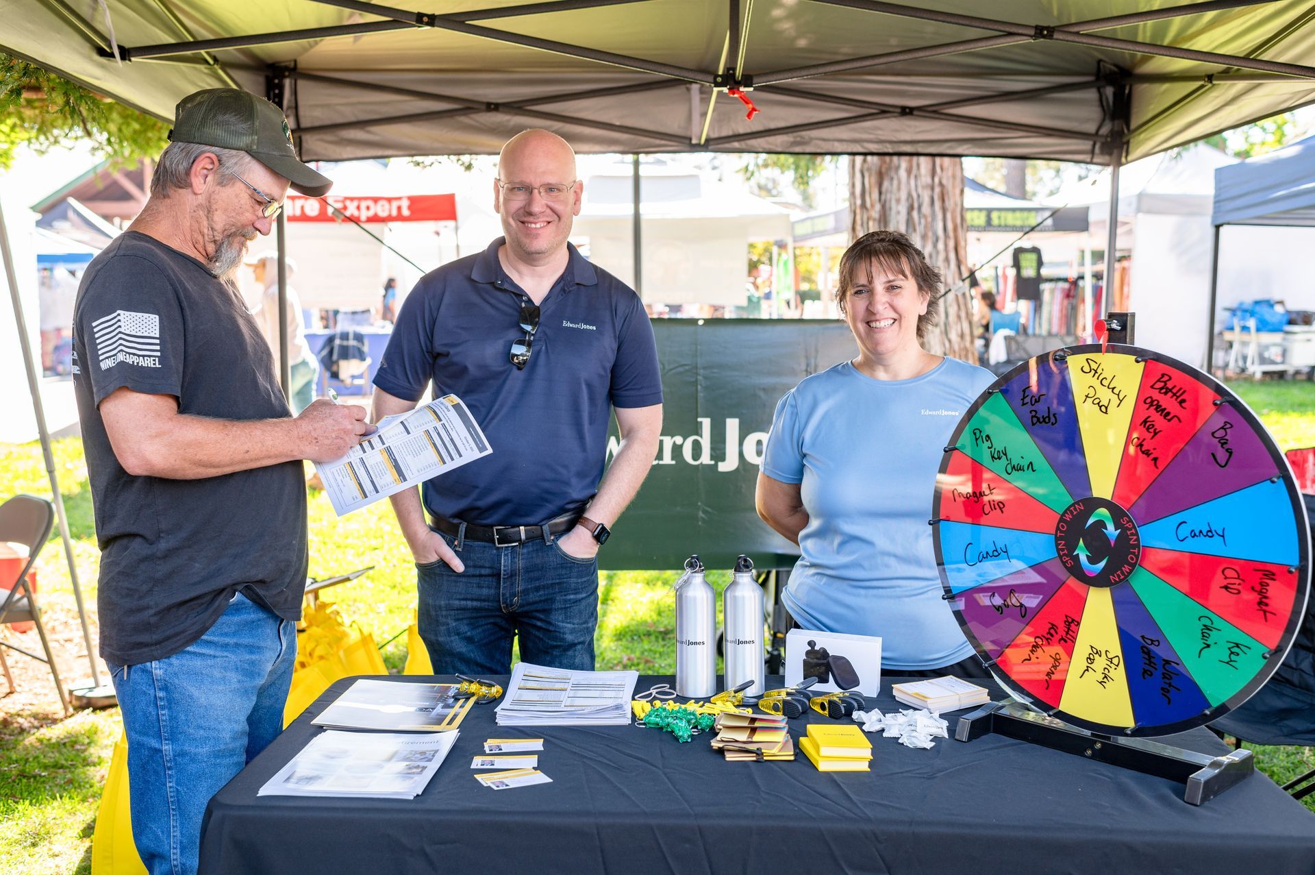 Three people at a table with flyers and a colorful prize wheel at an outdoor booth.