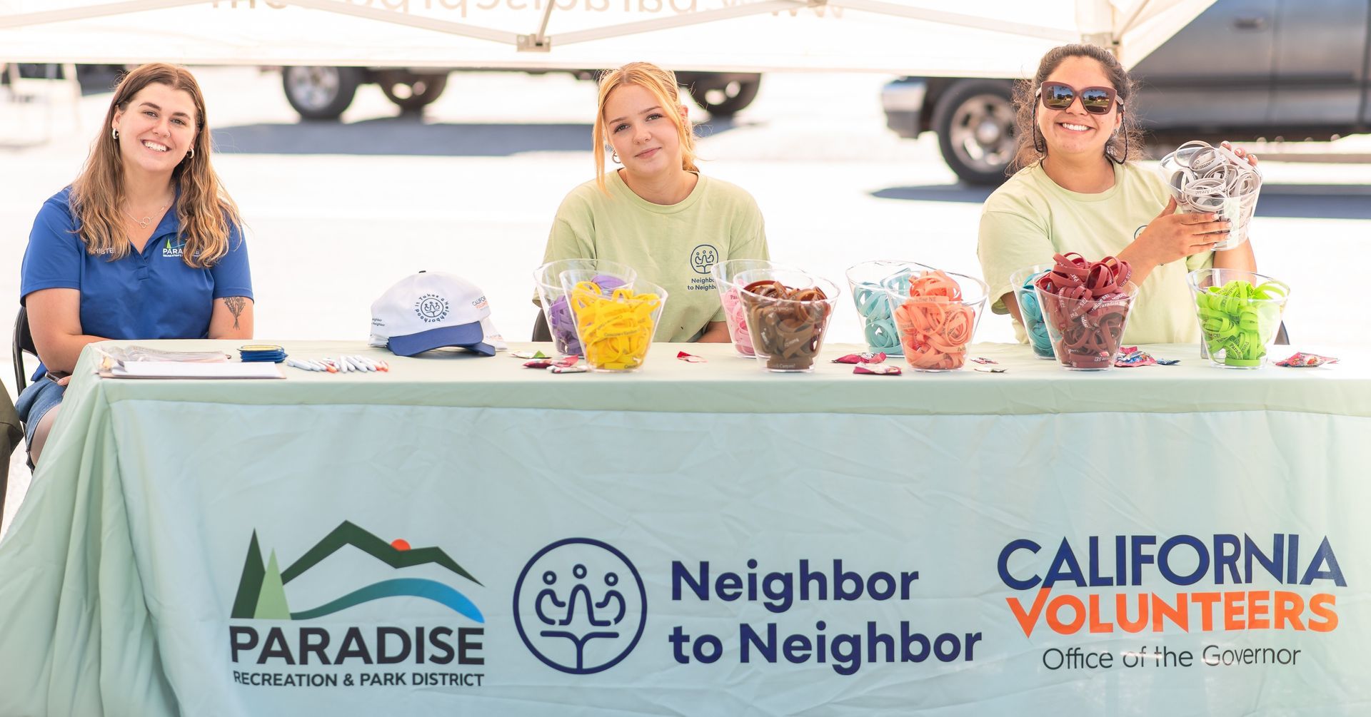 People behind a table with “Paradise,” “Neighbor to Neighbor,” and “California Volunteers” signs at an event