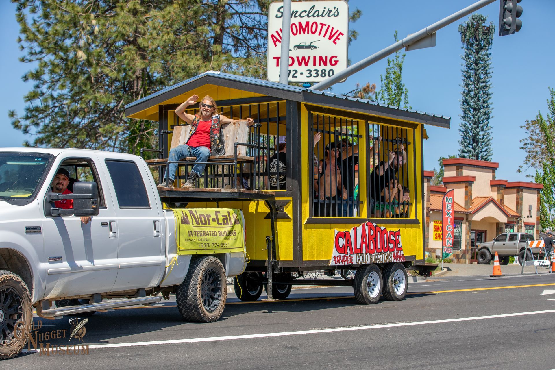 A white pickup truck pulls a yellow jail-themed float in a parade, featuring people in a barred cage.
