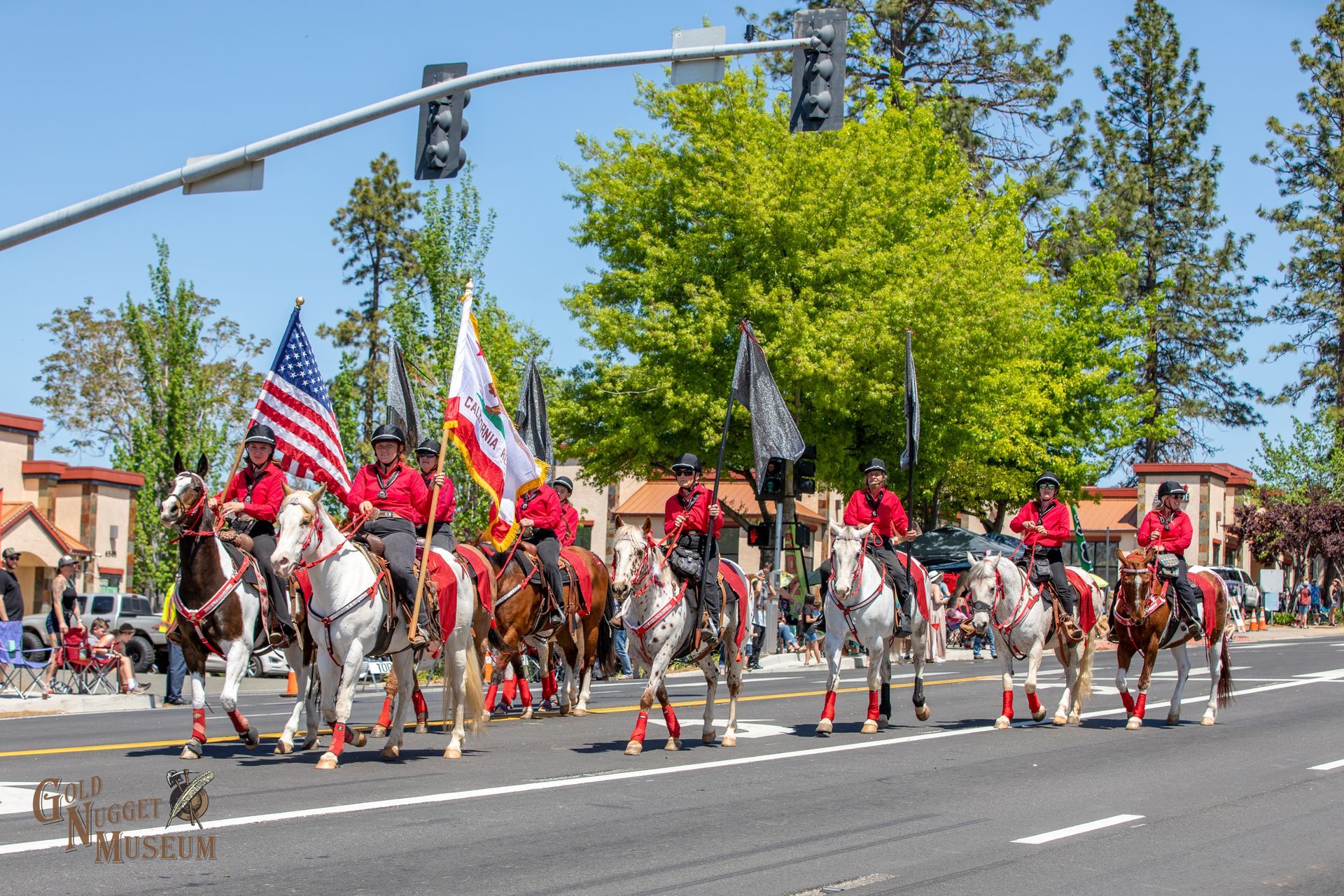 A mounted color guard rides horses down a street during a sunny outdoor parade, carrying American and state flags.