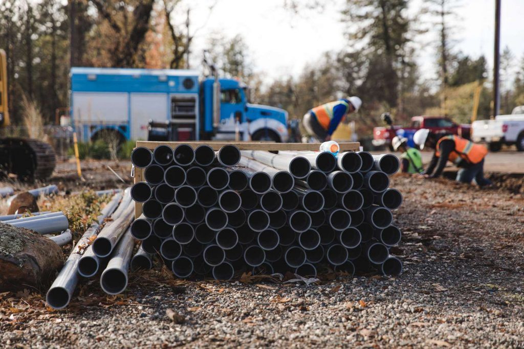 Stacks of black pipes at a construction site, with workers and a blue truck in the background.