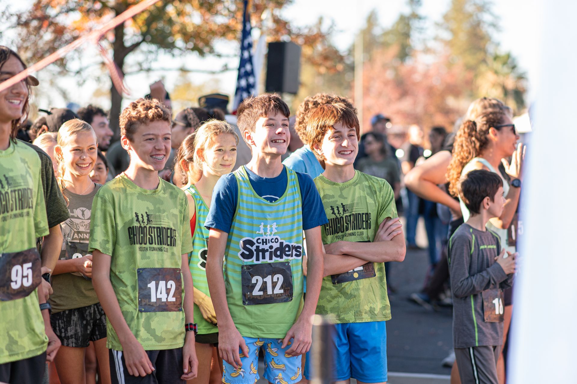 Cross-country runners in green shirts gather at the starting line outdoors under bright sunlight