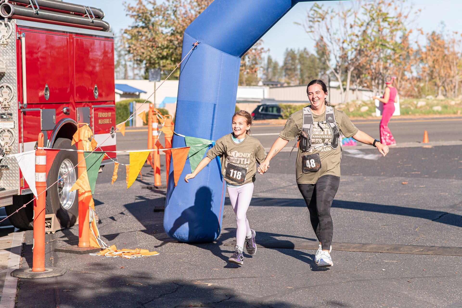 Children run under a blue inflatable arch during an outdoor race finish, with a red truck and cones nearby.