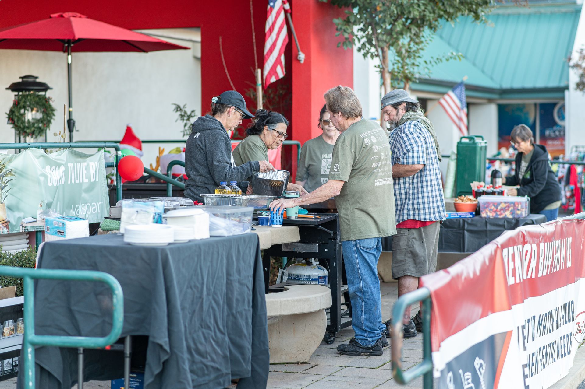 People at a market stall under a red umbrella, examining items on a table with a banner.