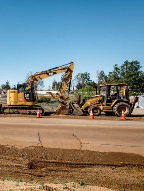 Yellow excavator loading debris into a backhoe on a roadside construction site under a clear blue sky