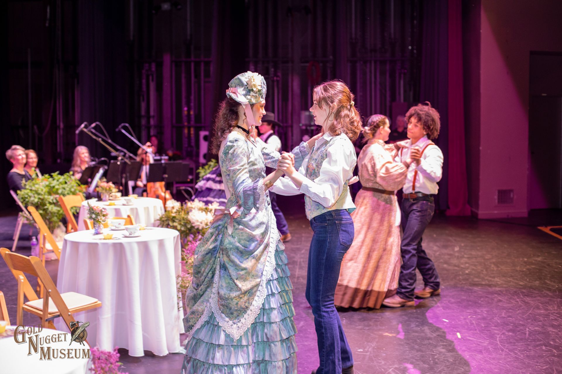 Two women dance on stage, a band plays in the background. Elaborate costumes, event setting.