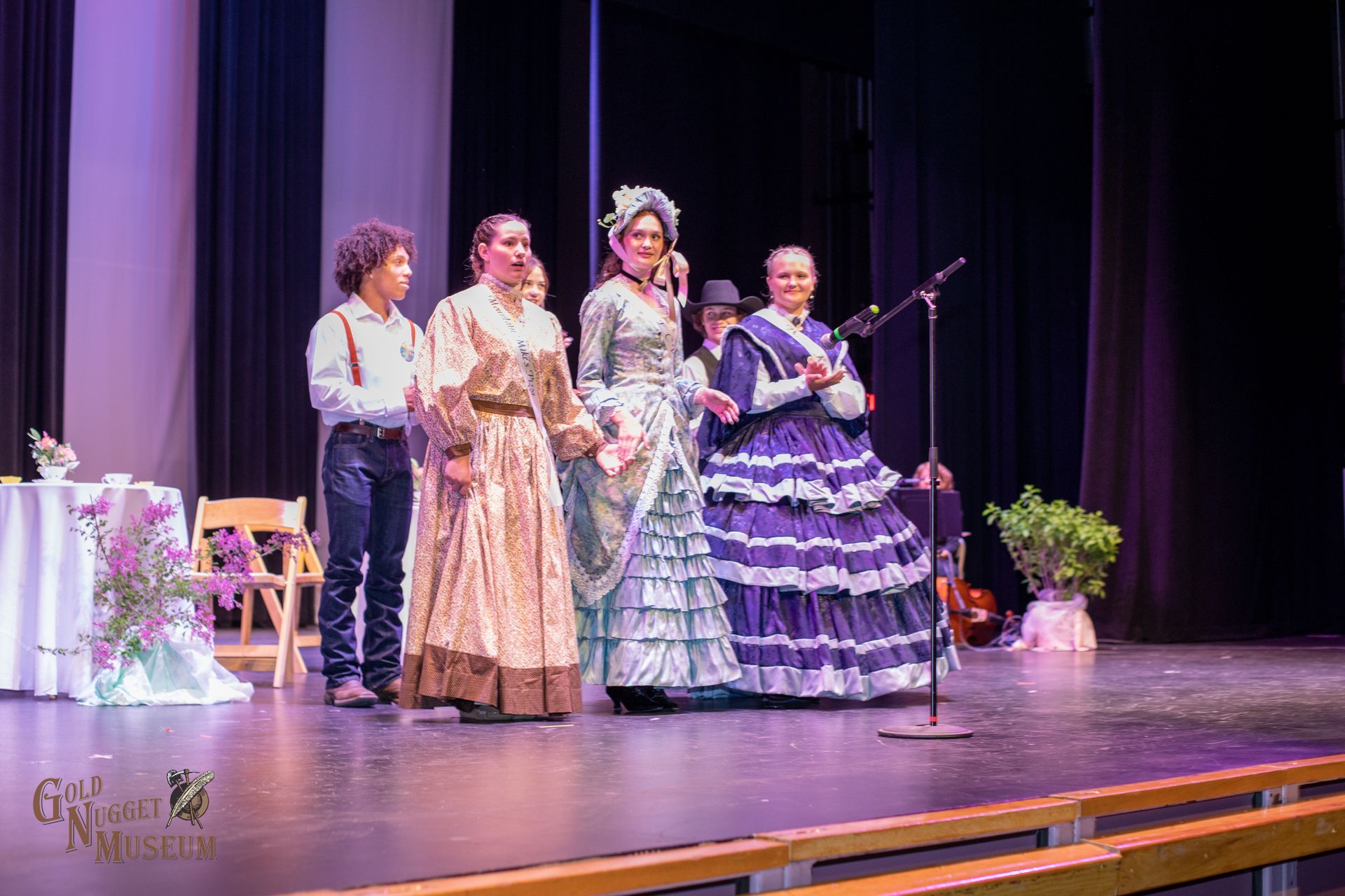 Actors in period costumes on stage, singing with microphones, at a theatre.