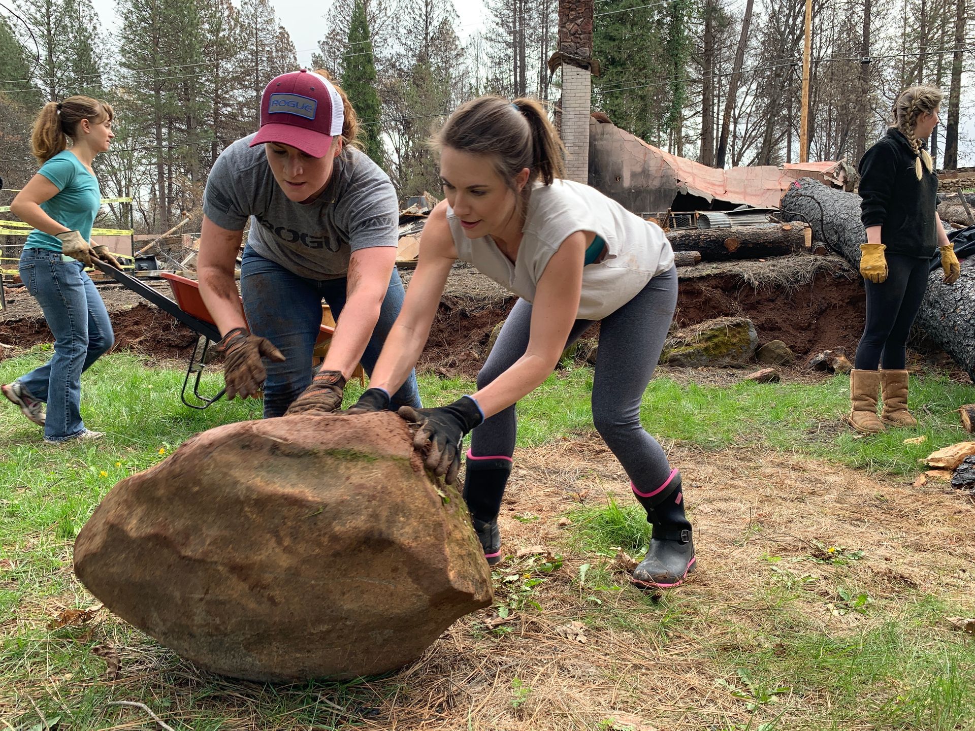 People lifting a large rock outdoors in a wooded area, with several others in the background