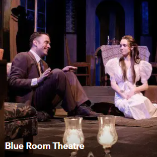 Two actors seated on a stage set, one in a suit and one in a white dress, with Blue Room Theatre text