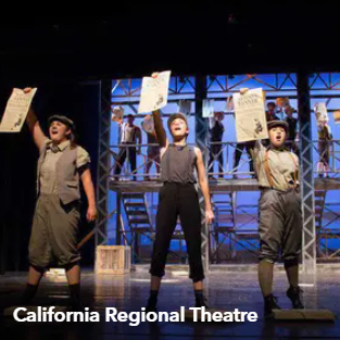 Three performers on a lit stage holding papers overhead, with scaffolding in the background.