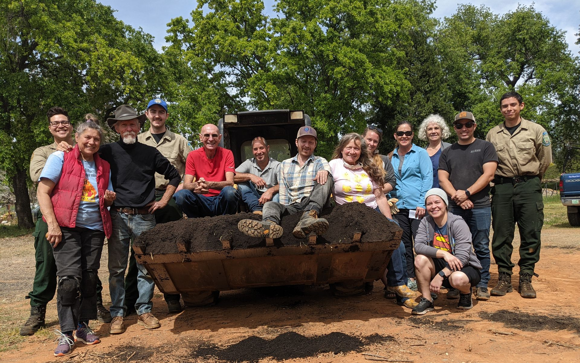 Group posing with a large muddy machine outdoors on a dirt lot, with trees in the background