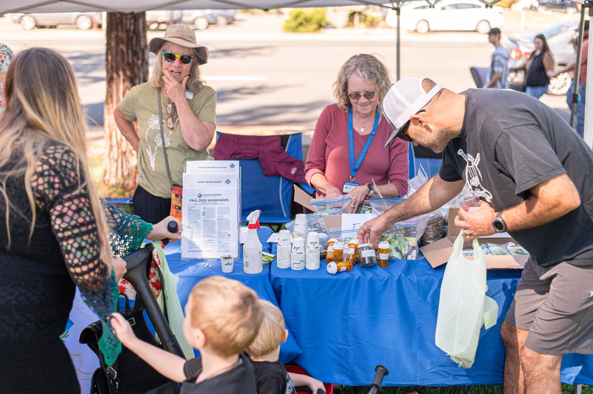 People at an outdoor market table with drinks, snacks, and a blue tablecloth