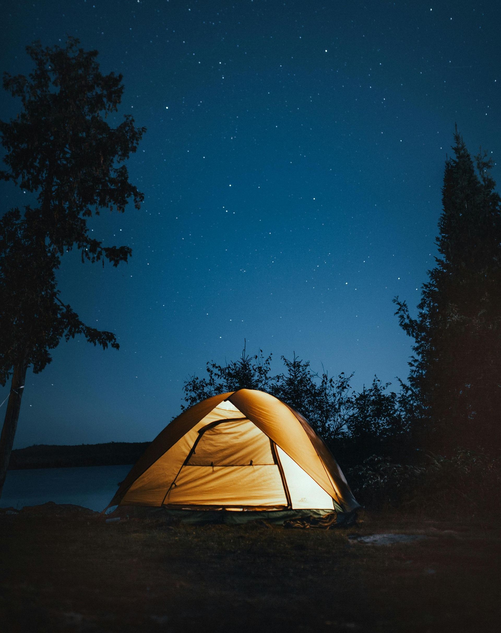 A lit camping tent under a dark blue starry sky, surrounded by trees near a body of water.