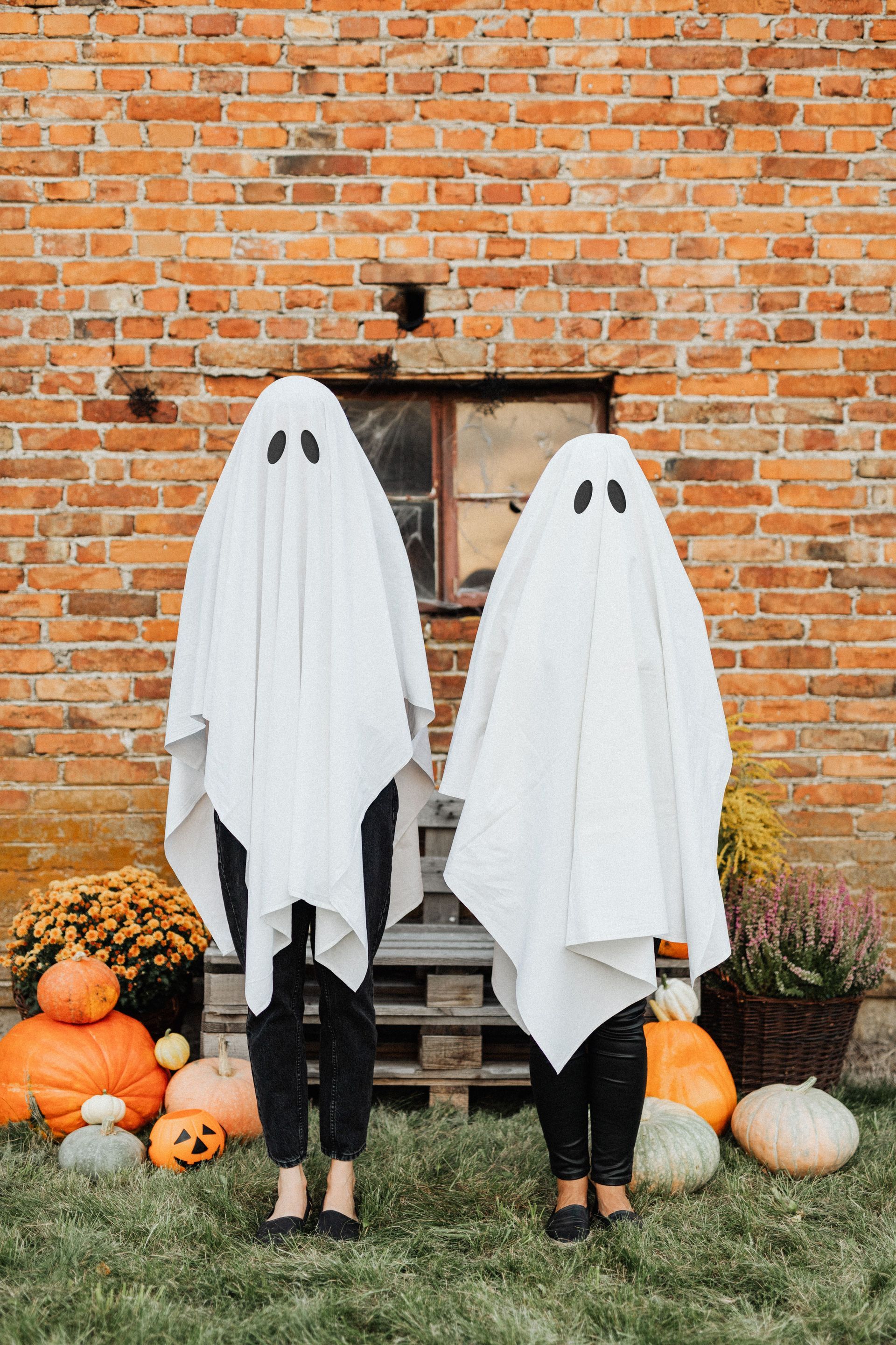 Two children in ghost costumes standing by pumpkins against a brick wall.